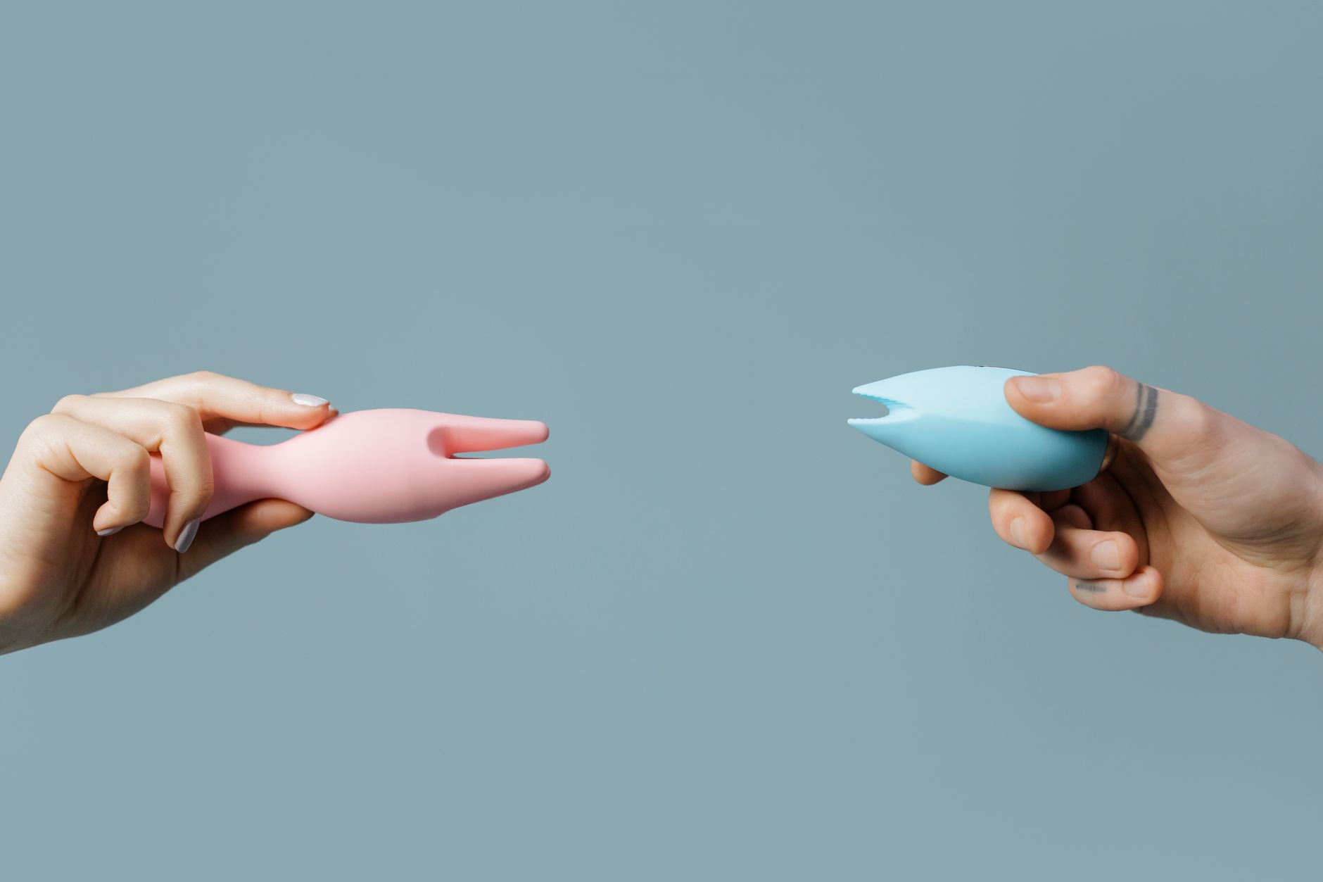 Duas mãos segurando vibradores de silicone rosa e azul em um fundo pastel