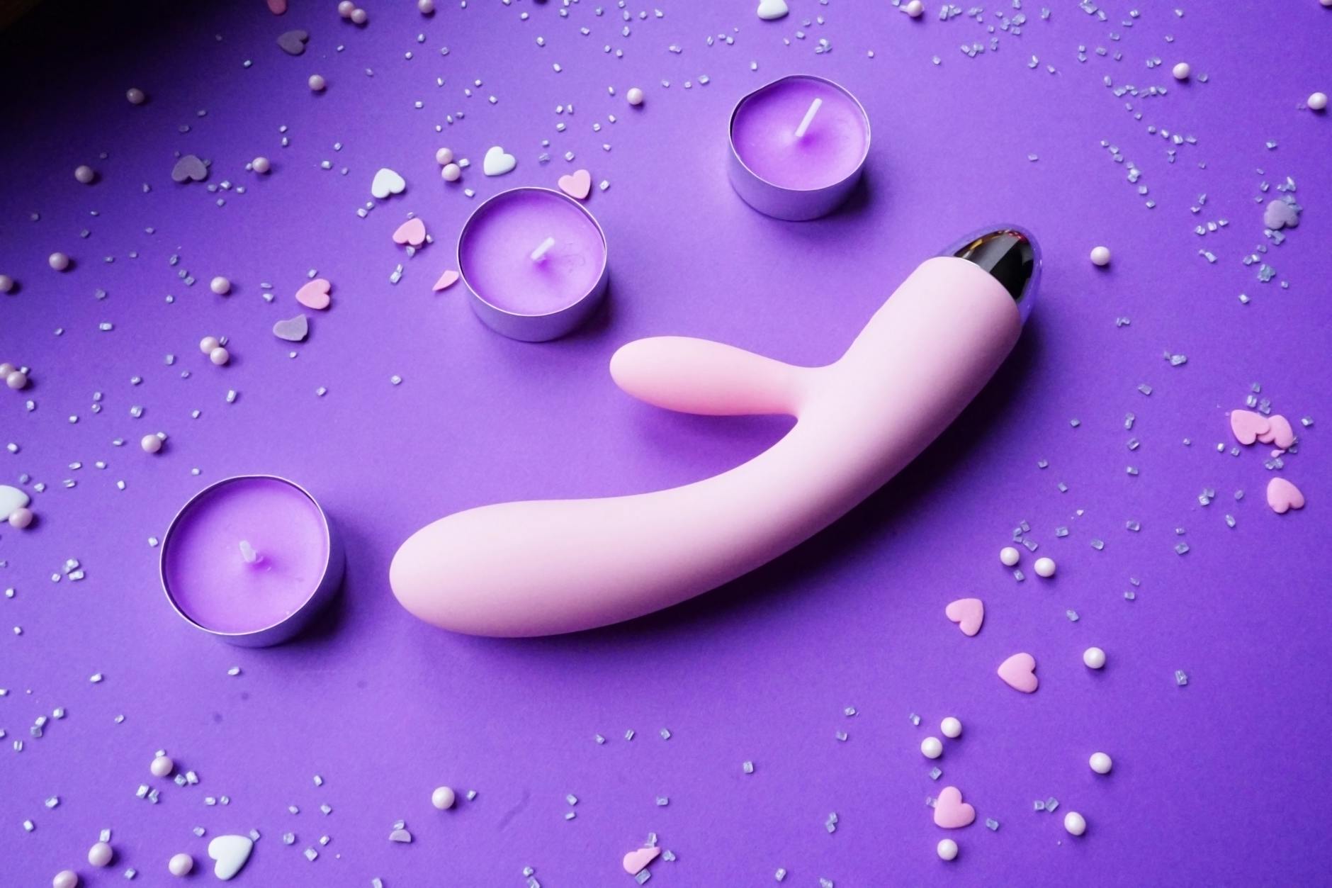 Vibrador rosa em fundo roxo com confete em formato de coração e velas para ambiente romántico